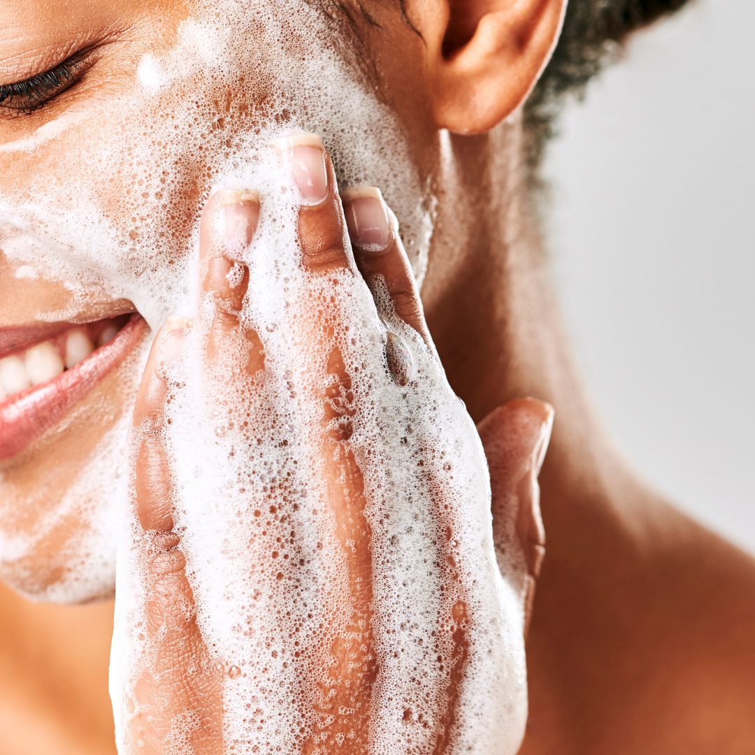 A smiling woman is cleansing her face with a super foamy facial cleanser.