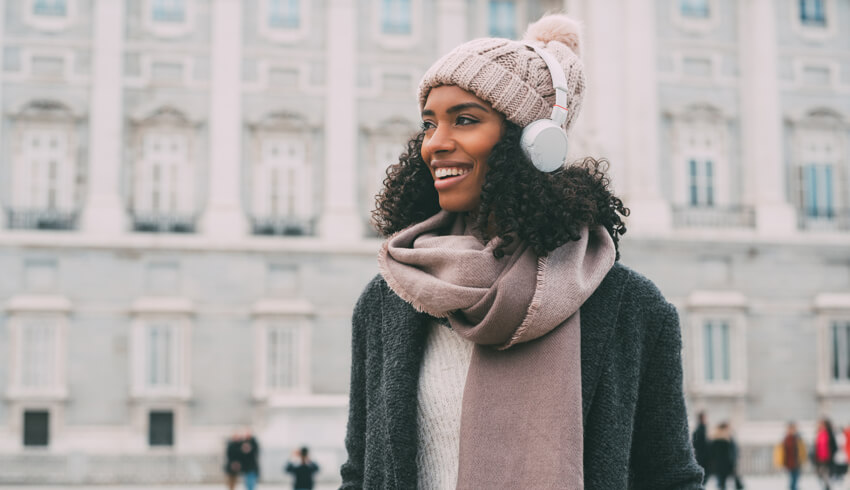 A smiling woman with glowing skin is standing in front of a building wearing a woollen jacket, large scarf, matching beanie and white headphones.